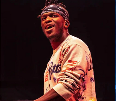 KSI wearing a patterned white long-sleeved shirt and bandana, smiling while performing on stage under warm lighting.