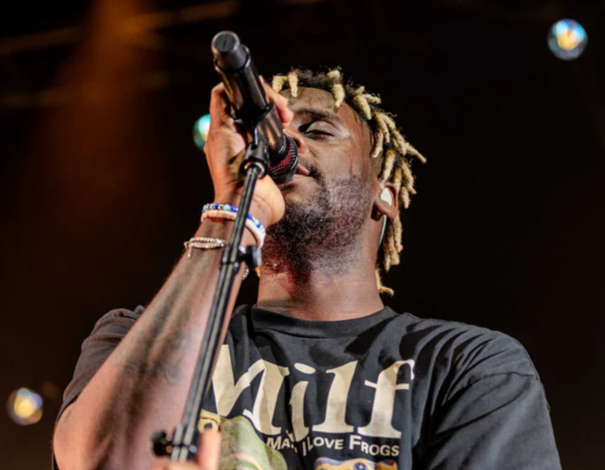 A person with blonde-tipped dreadlocks singing into a microphone on stage, wearing a t-shirt with text.