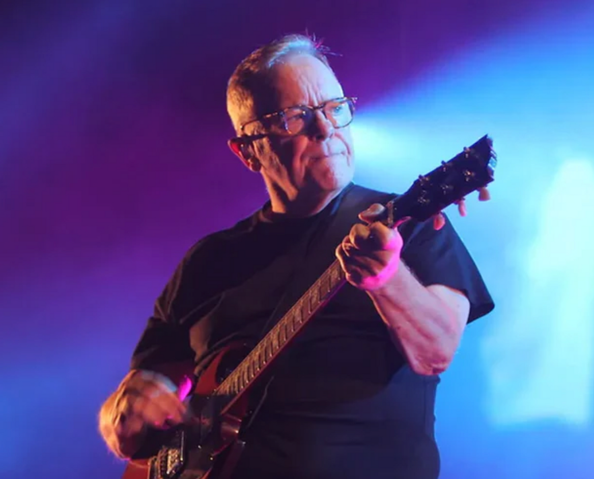 A person with glasses playing an electric guitar under blue and purple stage lighting.
