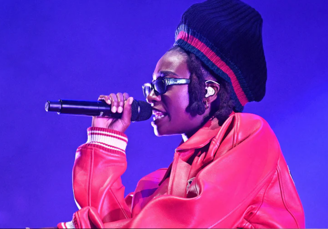 A performer in a red leather jacket and knit cap sings into a microphone against a vibrant purple stage background.