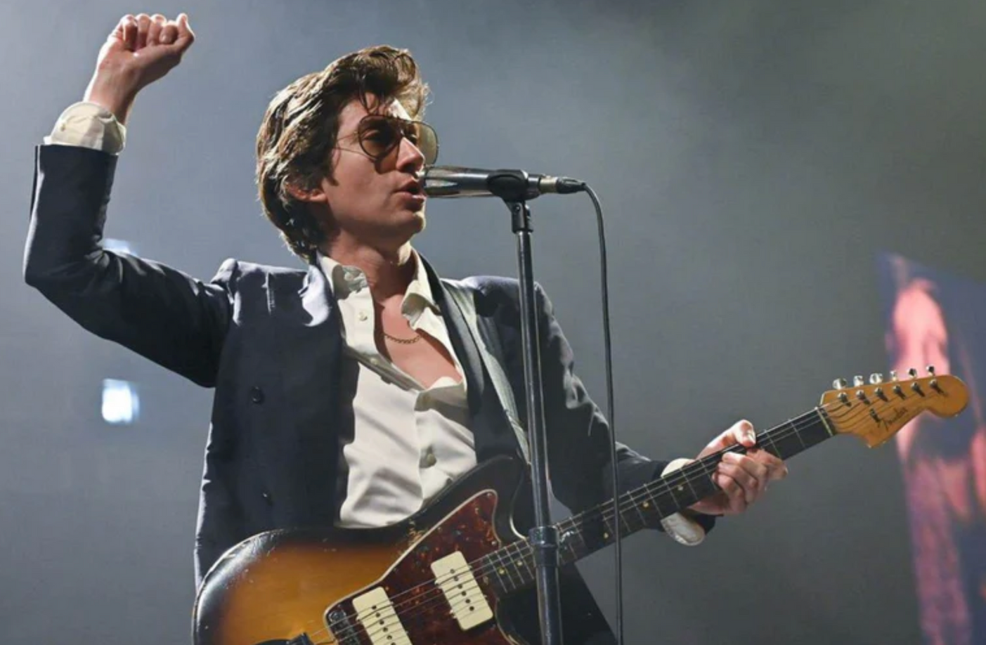 A musician in a suit and sunglasses singing into a microphone while playing an electric guitar under stage lighting.