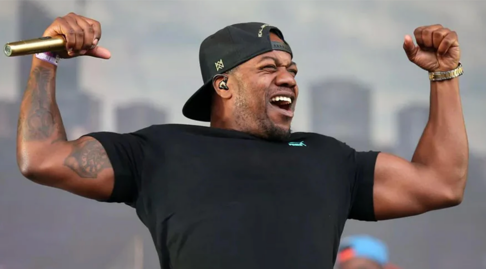 A smiling performer in a black t-shirt and cap raises both arms with a gold microphone in one hand during an outdoor show.