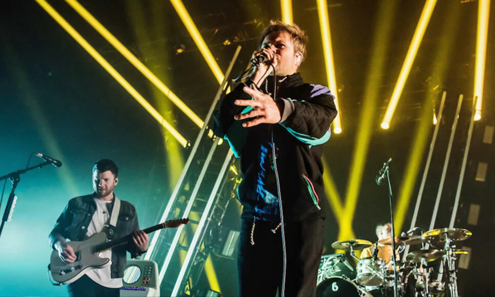 A lead singer performs into a microphone on stage, accompanied by a guitarist and drummer under bright yellow stage lights.