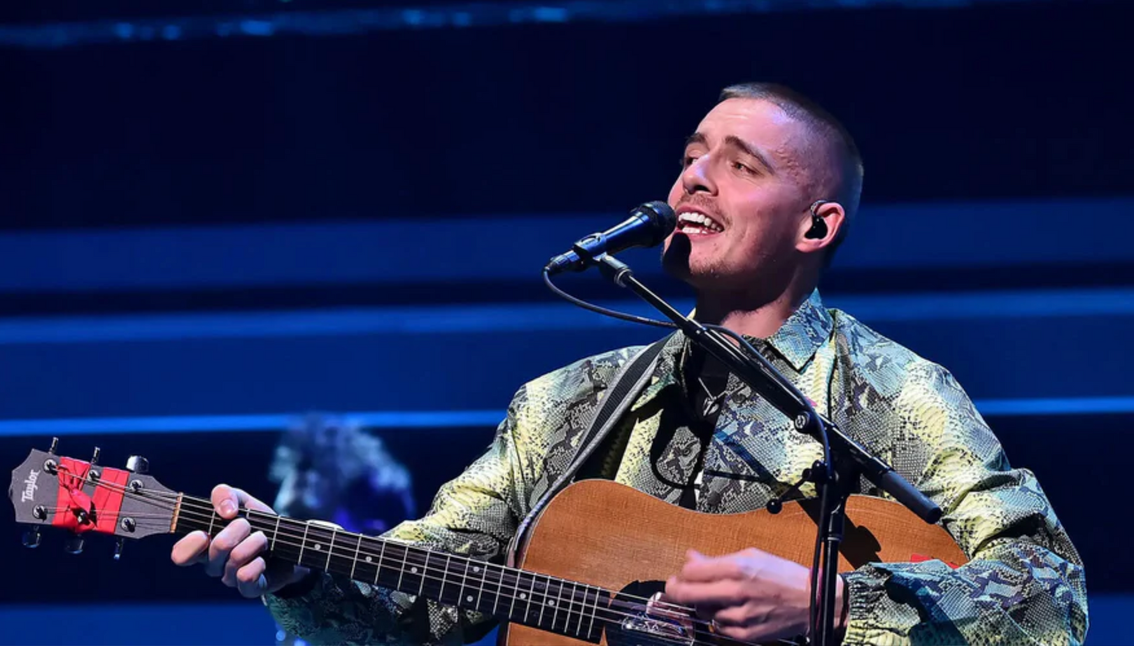 A singer in a patterned button-down shirt performing on stage while playing an acoustic guitar into a microphone.