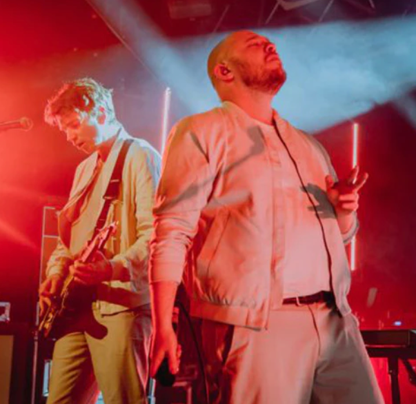 Two musicians perform on stage bathed in red light; one plays guitar while the other sings with eyes closed.