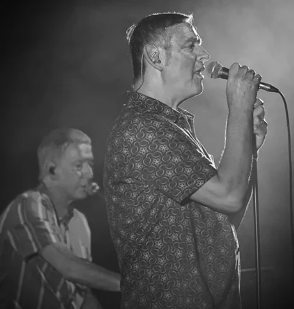 A singer in a patterned shirt performs at a microphone, with a musician playing keys in the blurred background.