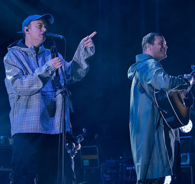 Two musicians perform on stage; one in a blue cap points while singing, the other plays an acoustic guitar.