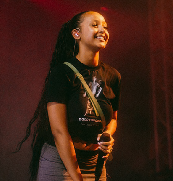 A person smiling on stage, wearing a black Paco Rabanne t-shirt and holding a microphone under warm lighting.
