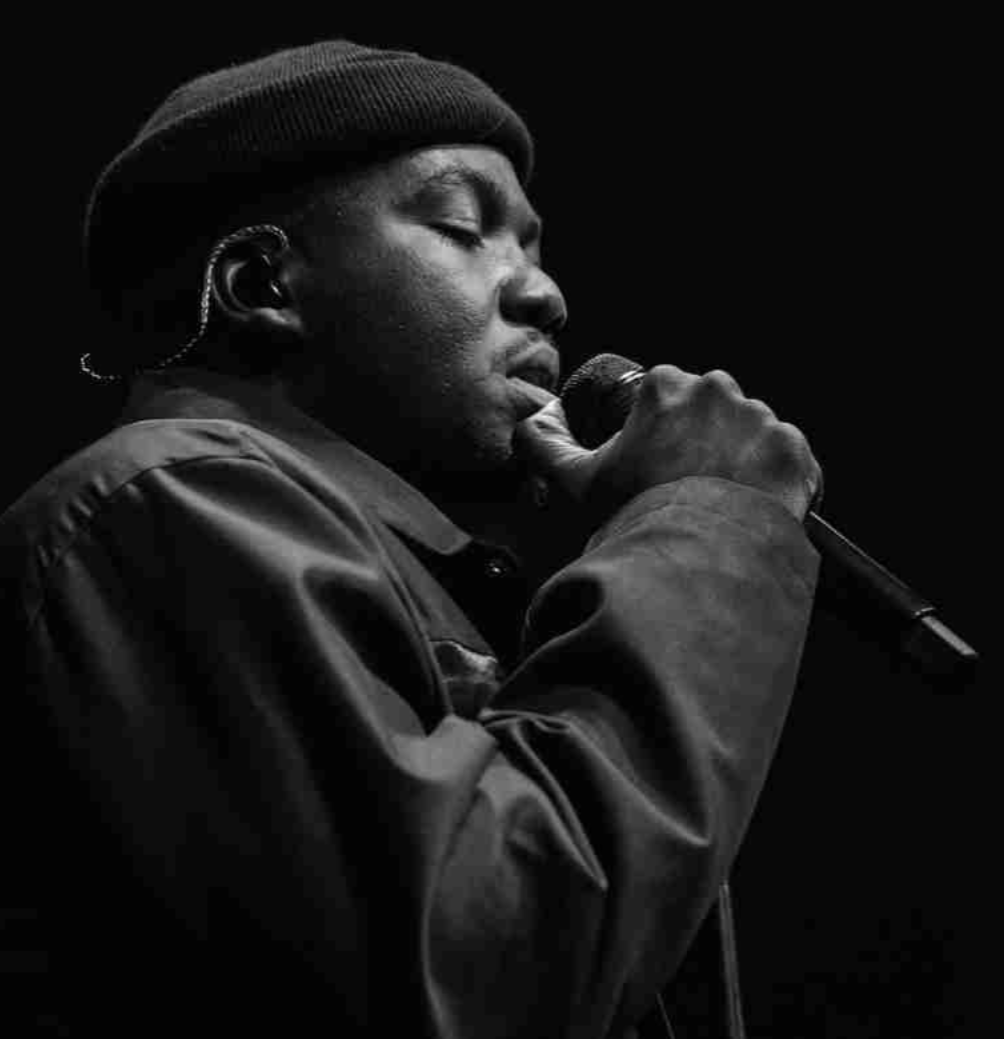 A musician in a beanie singing intensely into a microphone, captured in a black-and-white close-up shot.