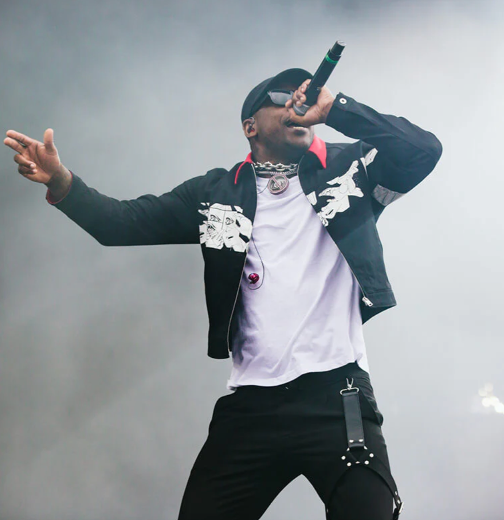 A performer in a black jacket and white t-shirt sings into a microphone on stage against a smoky background.