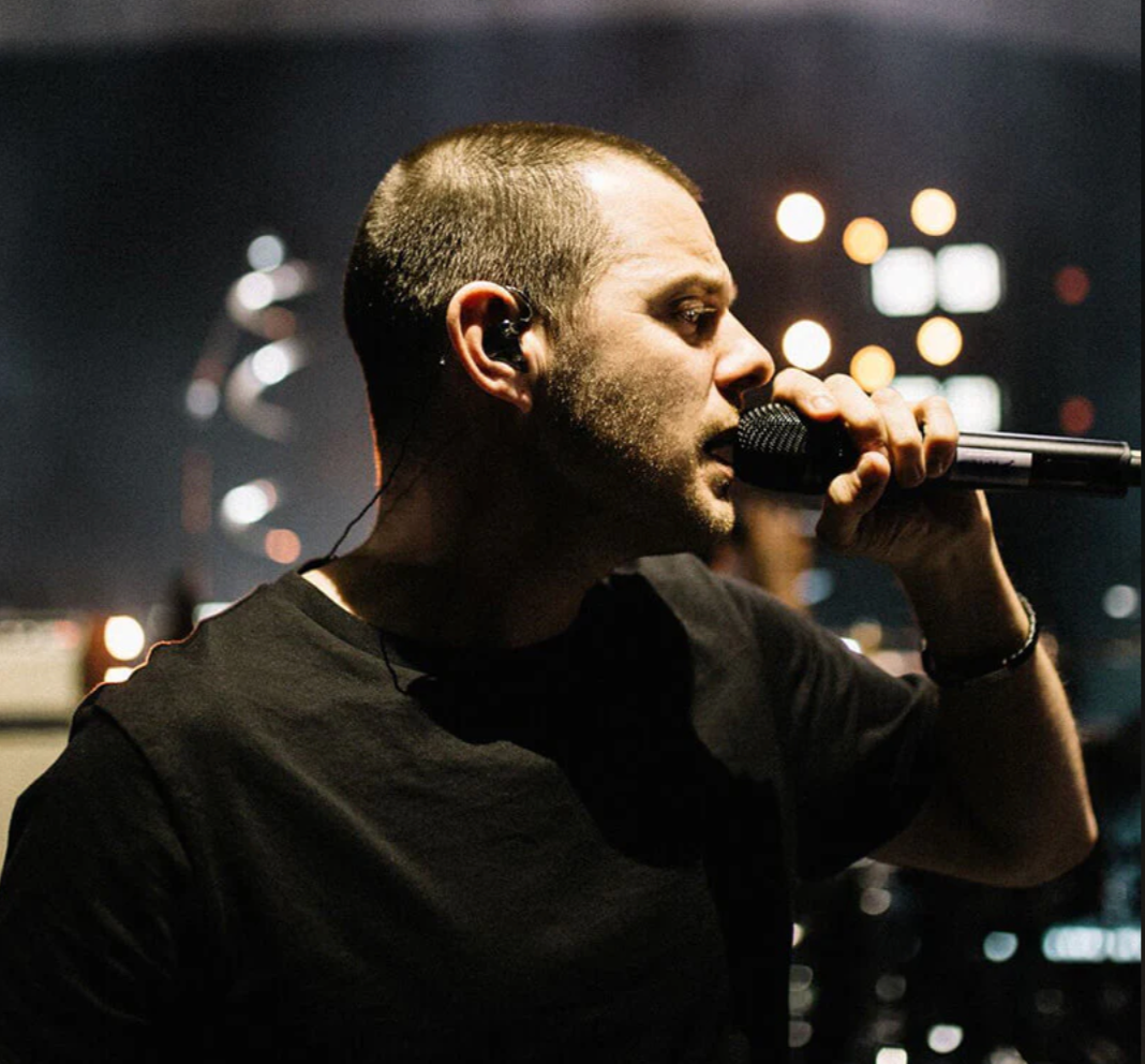 A man in a black t-shirt singing into a microphone on stage against a dark background with blurred stage lights.