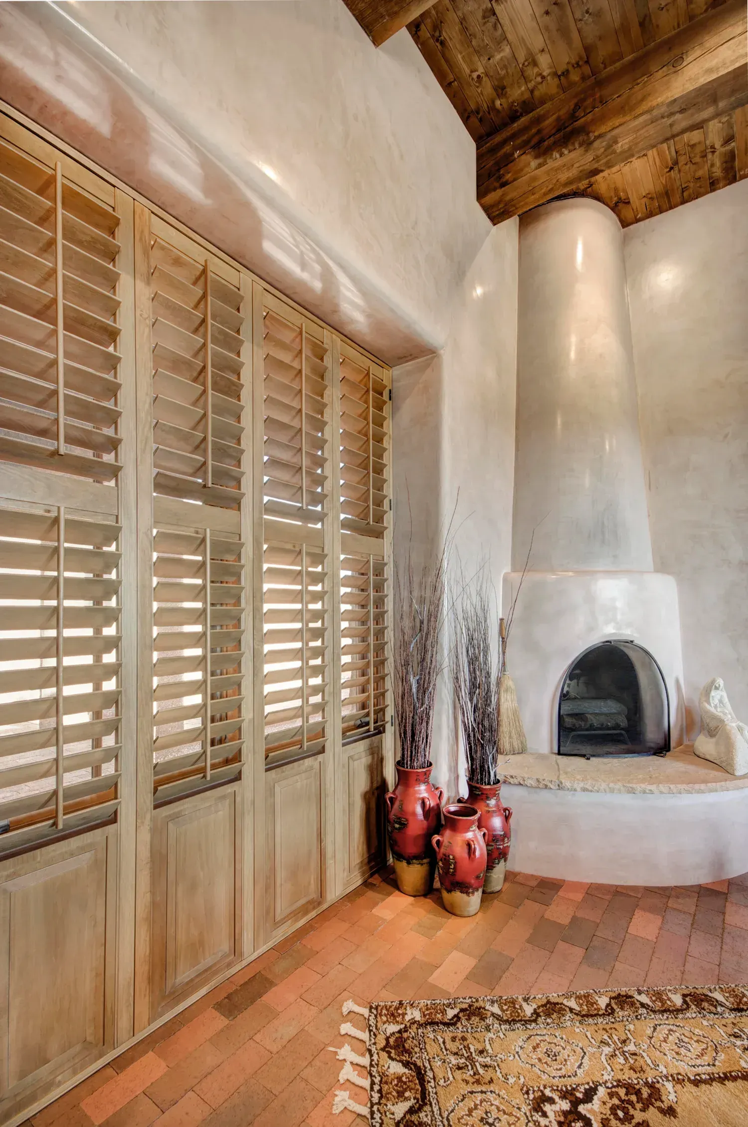 Rustic living room with wood shutters, a white kiva fireplace, terracotta floor, and decorative pottery.