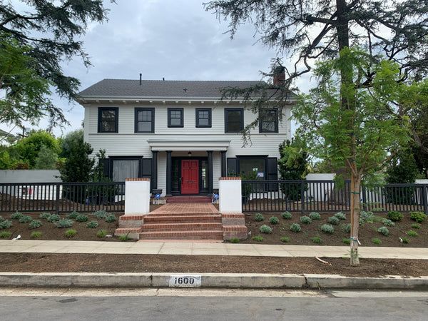Two-story white house with black shutters and a red front door, black fence, brick walkway, and freshly planted landscaping.