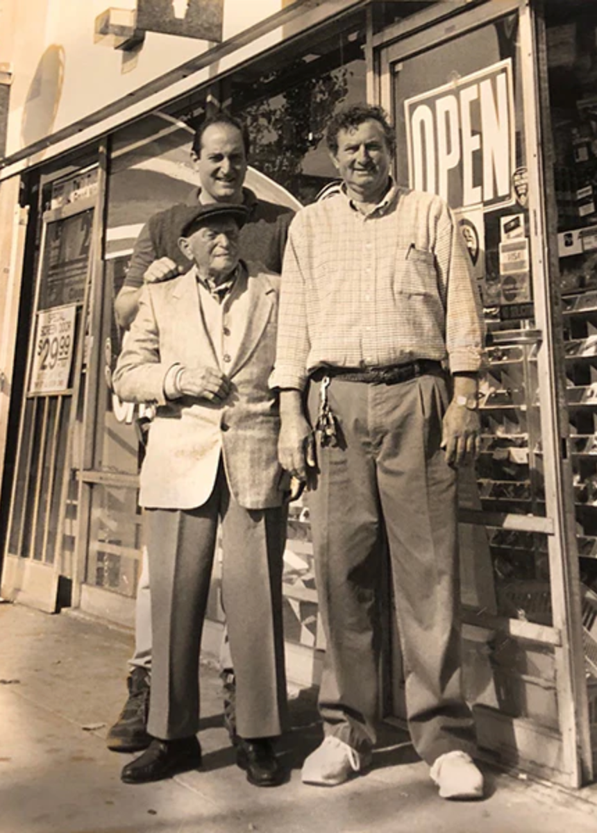 Three men pose outside a store; older man in front, two taller men behind. Storefront with