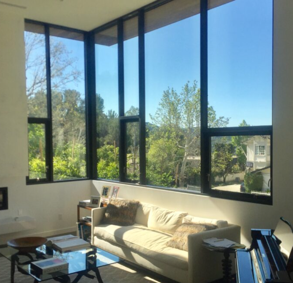 Living room with large windows overlooking trees and a sunny sky; couch, piano.