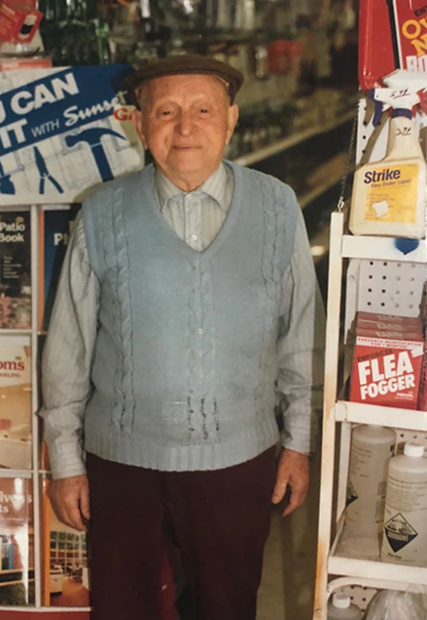 Elderly man in blue sweater and cap stands inside a store, a shelf of cleaning supplies to the right.