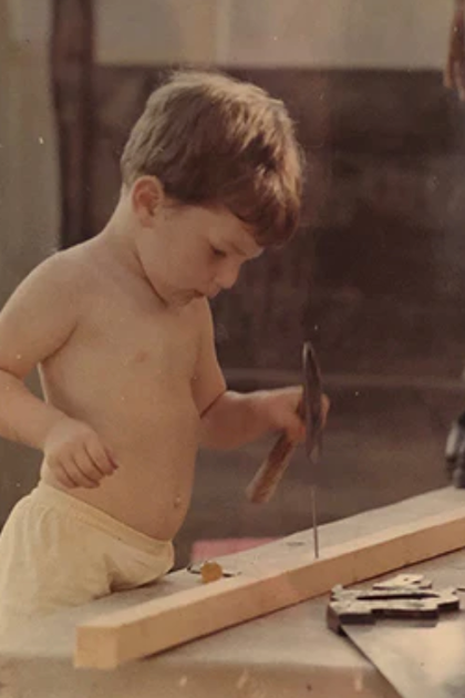 Young child hammering a nail into a wooden plank. Bare-chested, wearing white underwear, focused.