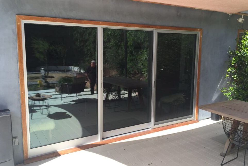 Sliding glass doors with wood trim reflect an outdoor patio with a person, table, and greenery.