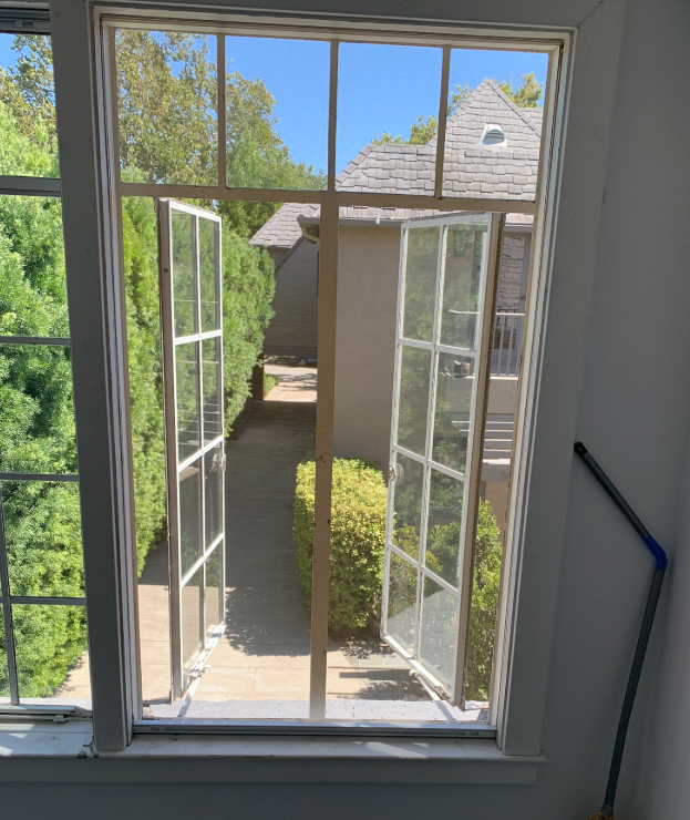Open, white-framed window with two panels ajar, overlooking a pathway with green foliage and a beige building.
