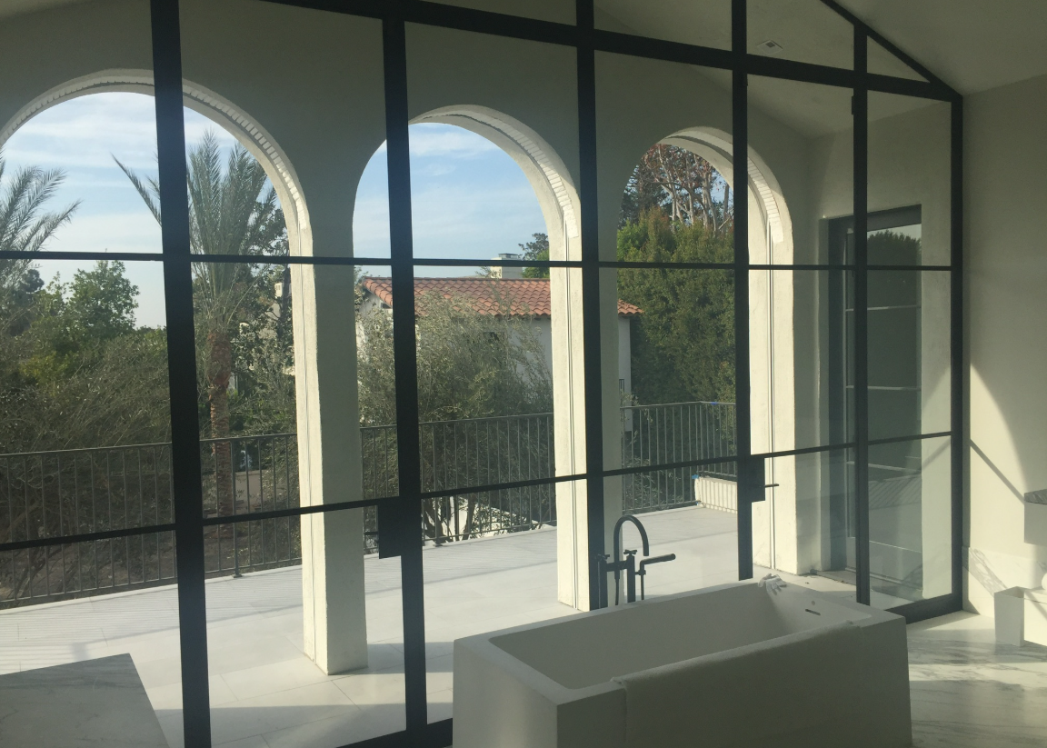 Bathroom with black-framed windows, white tub, and arched columns overlooking a green, sunny outdoor view.