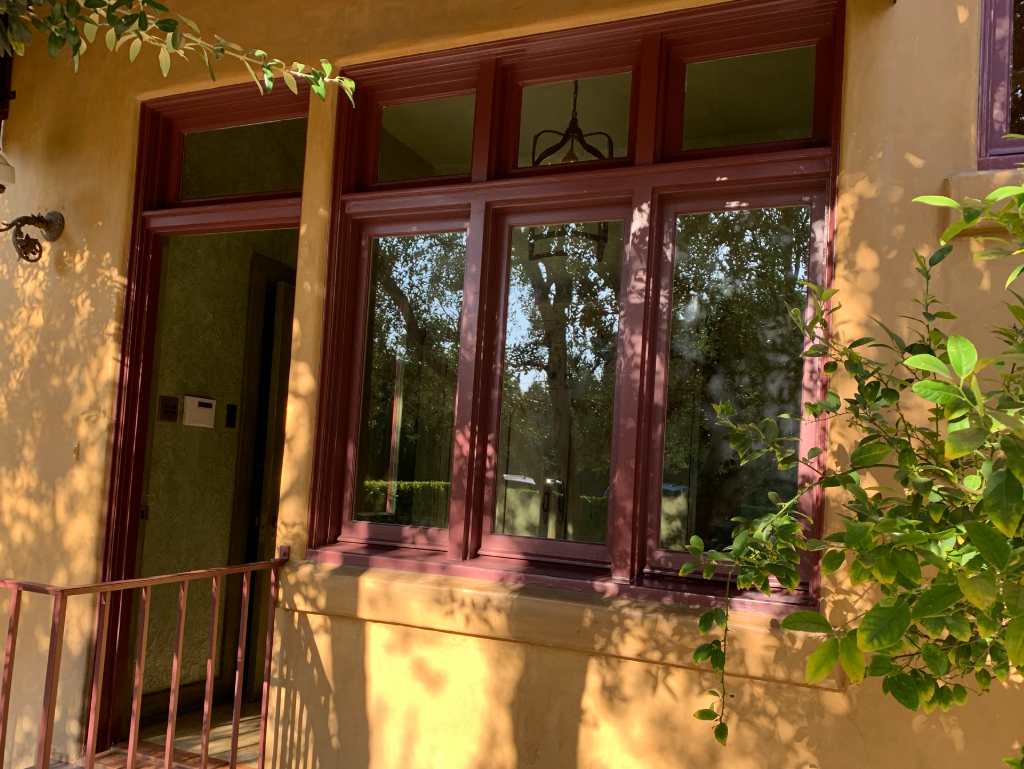 Window with burgundy trim reflecting trees, set in a yellow stucco wall.