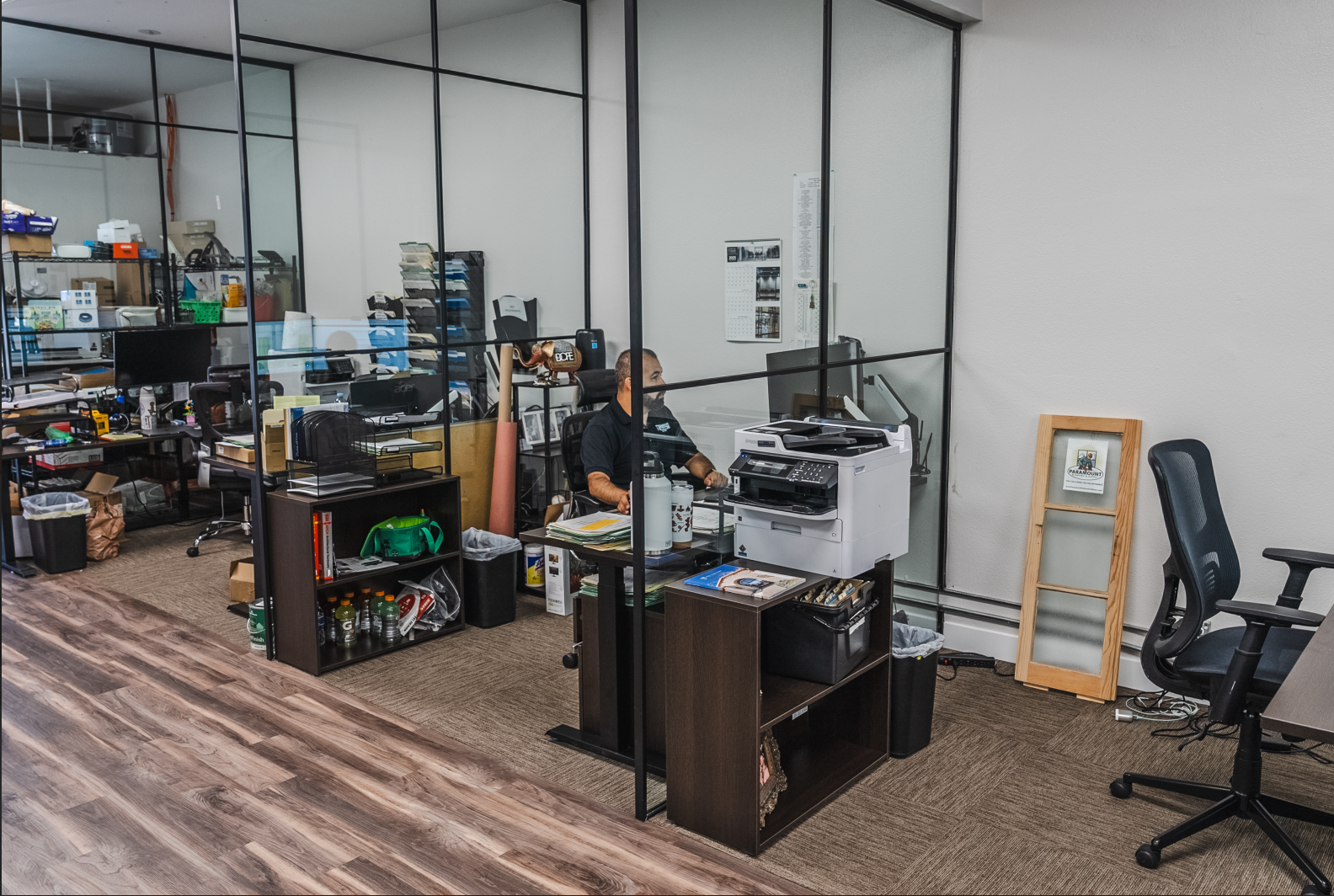 Office with person at a desk, printer, shelves with items, and black framed glass walls.