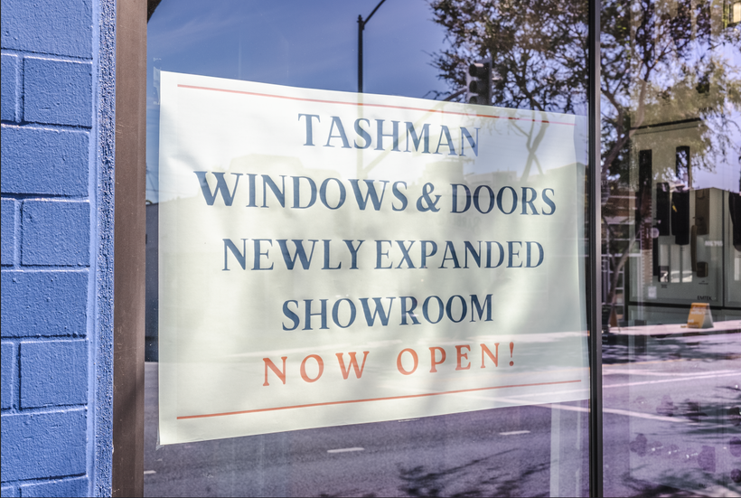 Sign in window of "Tashman Windows & Doors" announces a newly expanded showroom, now open, on a blue-painted building.