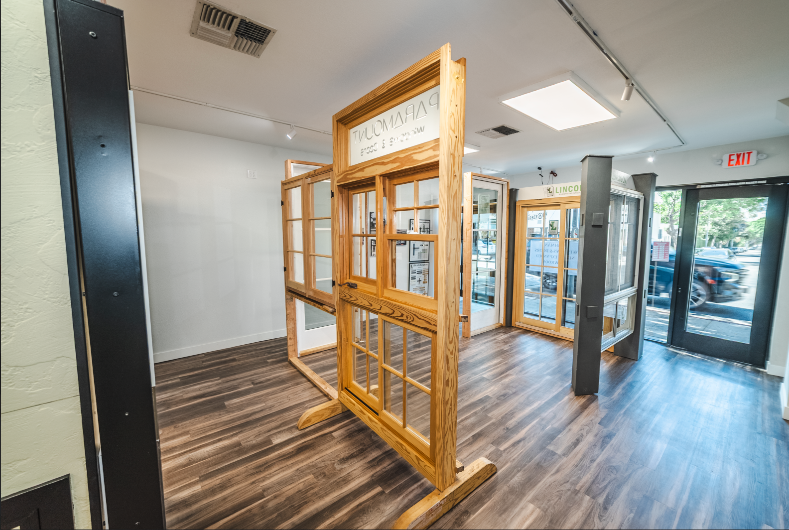Showroom with wood-framed windows and doors on display. Brown flooring, white walls, and a front door with a car visible outside.