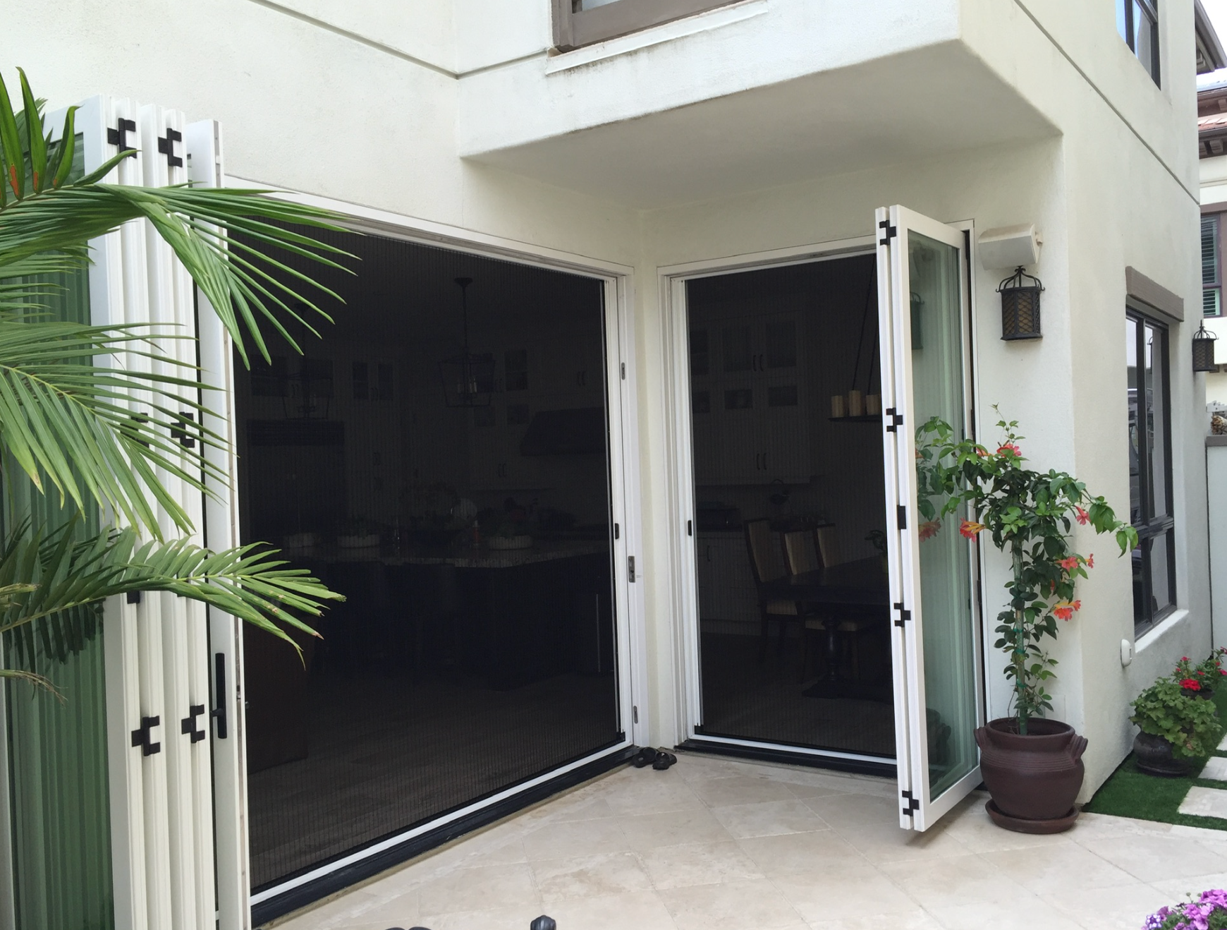 Folding glass doors open to a patio, palm frond on the left, potted plant to the right, white building.