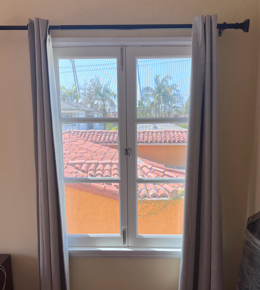 Window with gray curtains, overlooking red-tiled roofs and palm trees, beige walls.