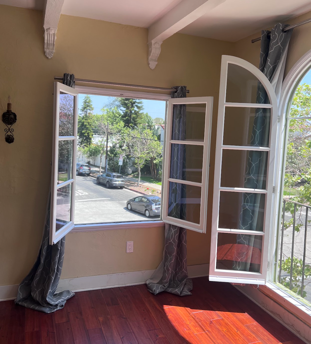 Open windows with flowing blue curtains, overlooking a street with parked cars and trees.