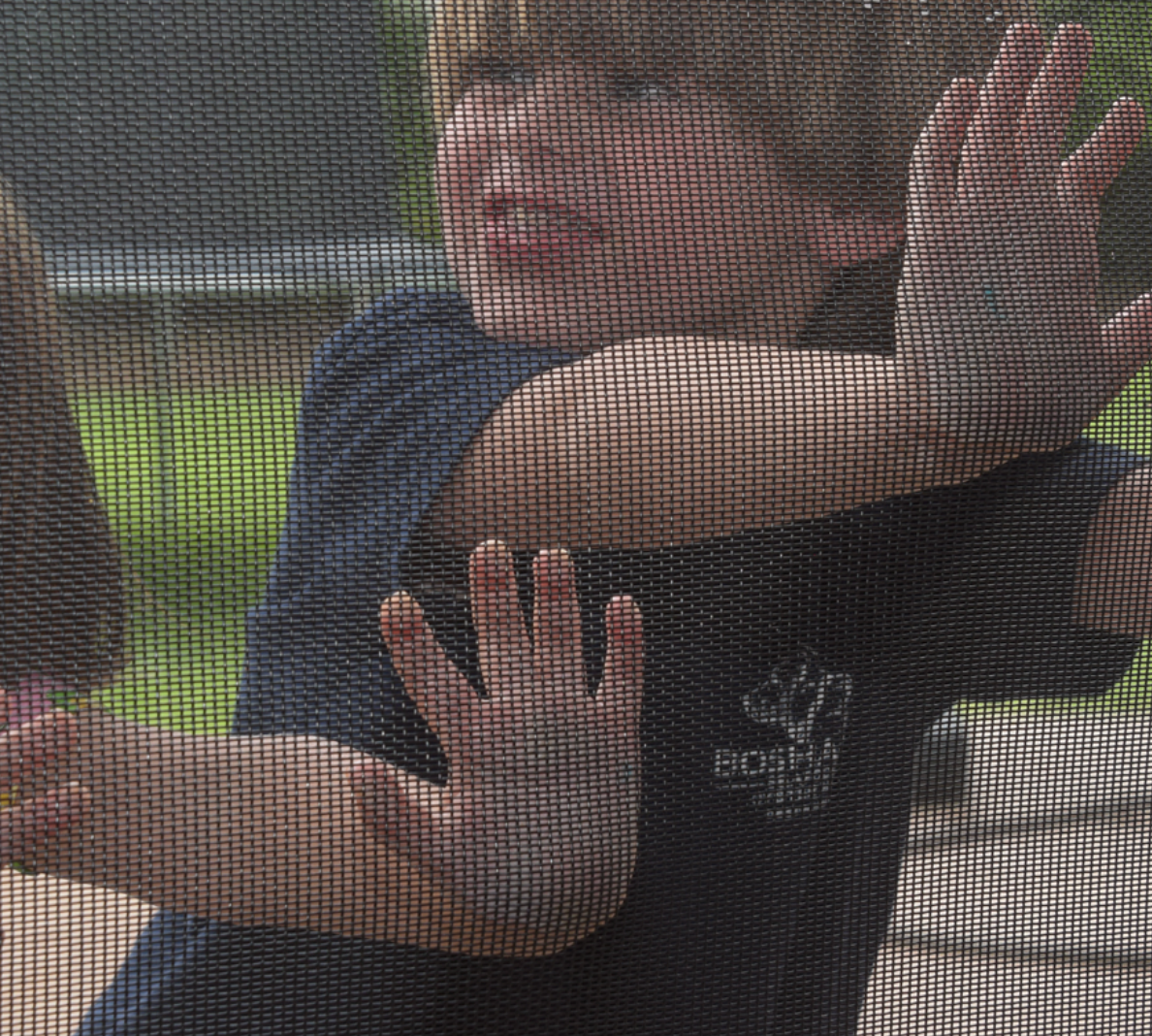 Boy pressing hands against a screen; green background, dark shirt, face visible through the mesh.