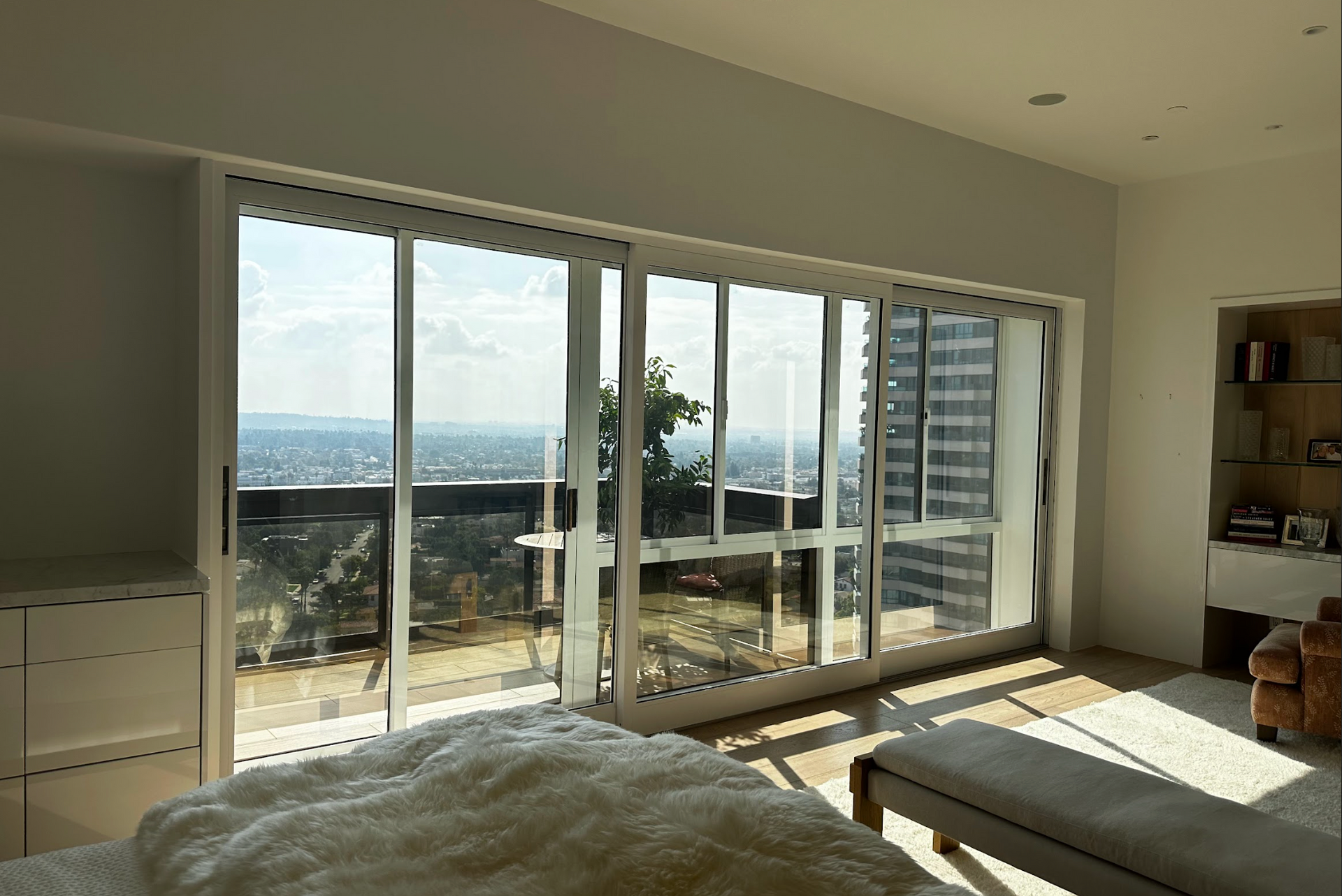 Bright bedroom with large windows overlooking a city, sunlight streaming in, bed in the foreground.