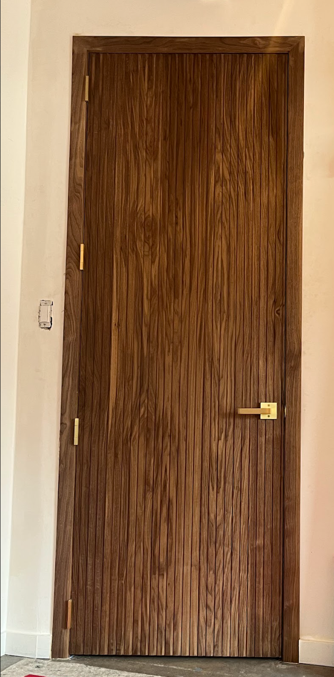 A wooden door with vertical grooves, gold hardware, and set against a white wall.