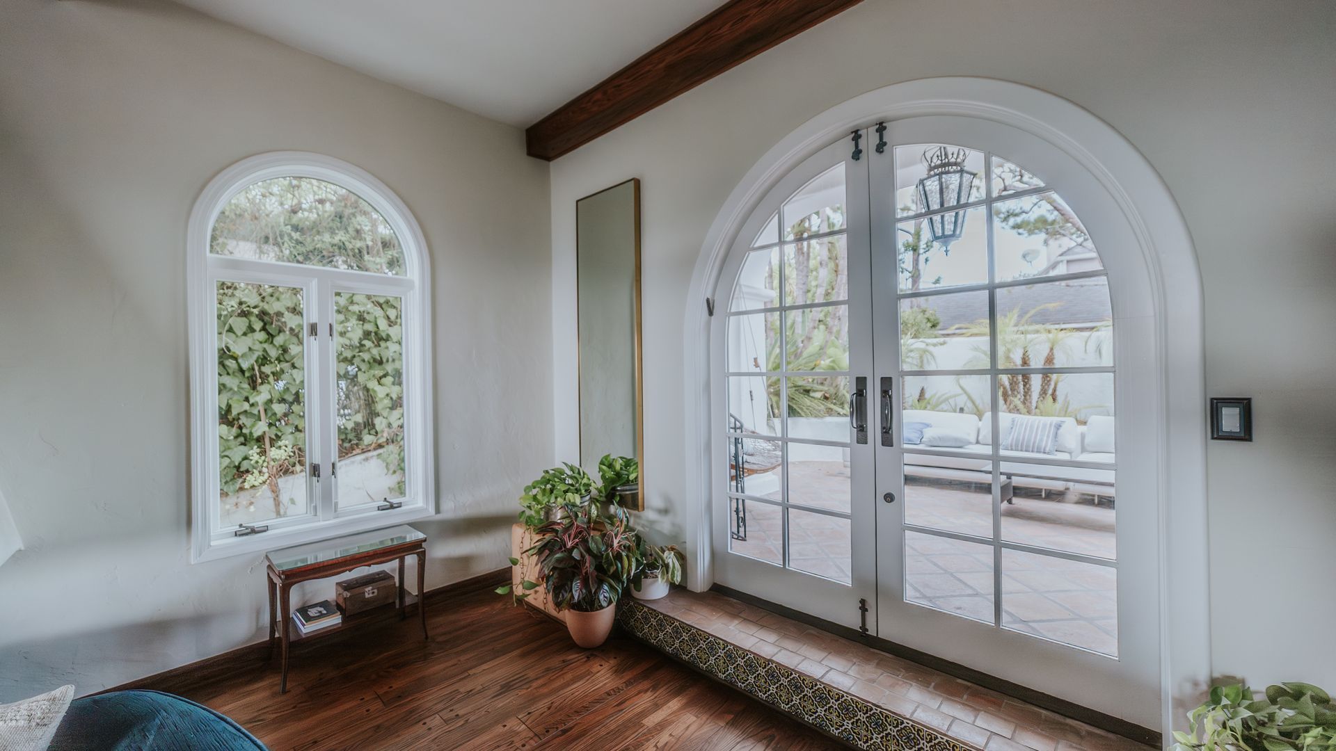 Arched white French doors open to a patio. Arched window, wood floors, and plants are visible.