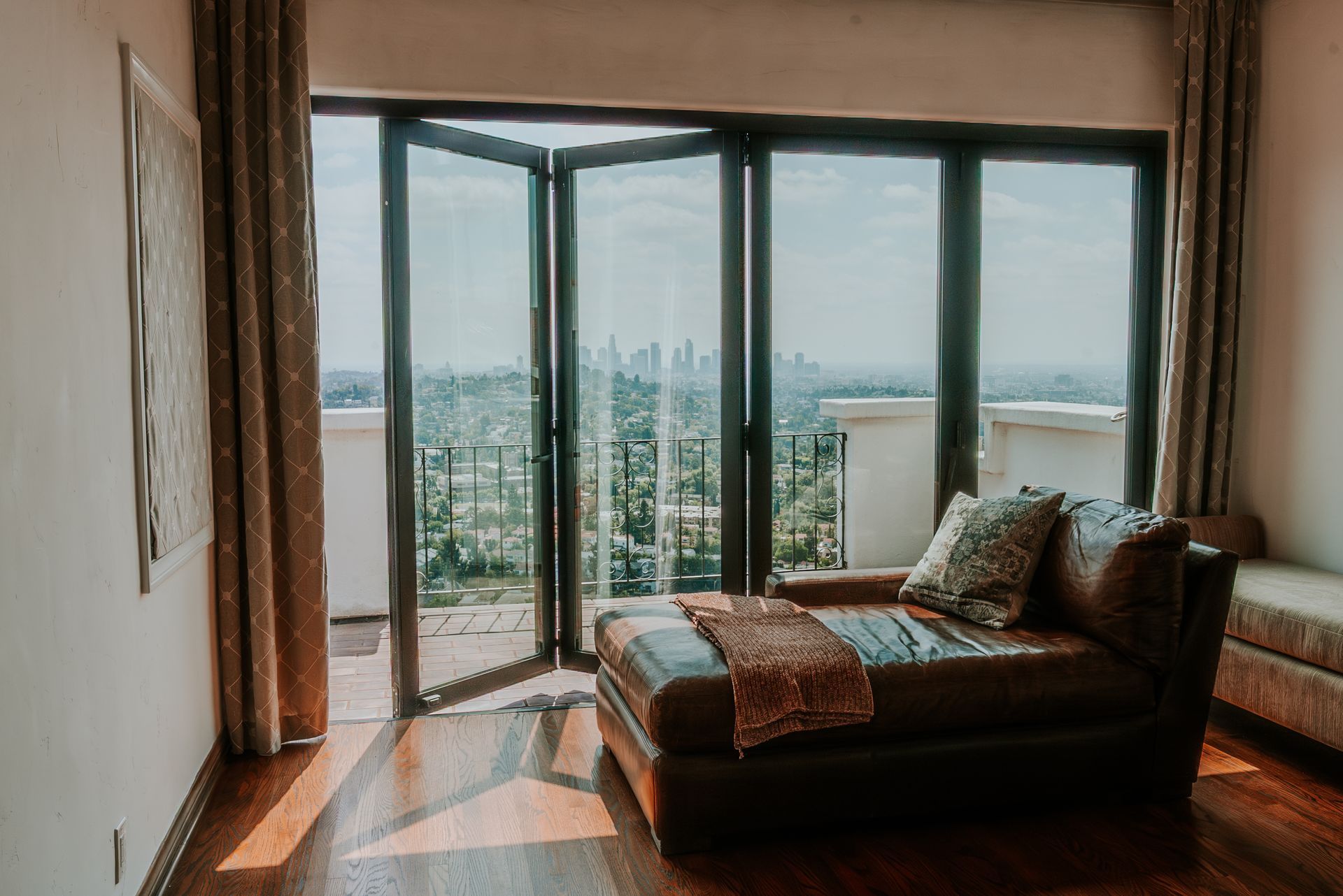 A room with a leather chaise lounge by open glass doors overlooking a city skyline, lit by sunlight.