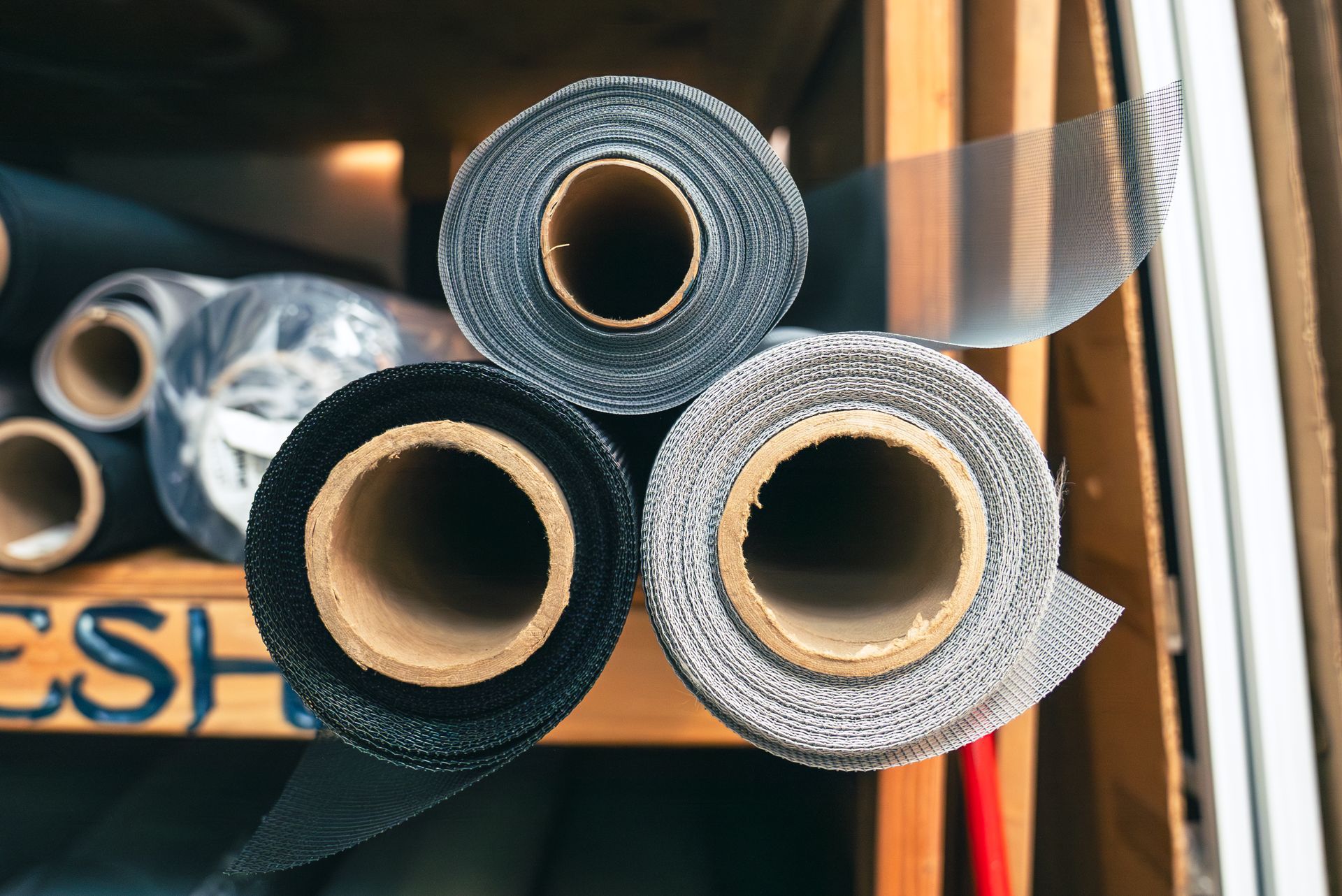 Three rolls of screen material on a wooden shelf: black, gray, and silver.