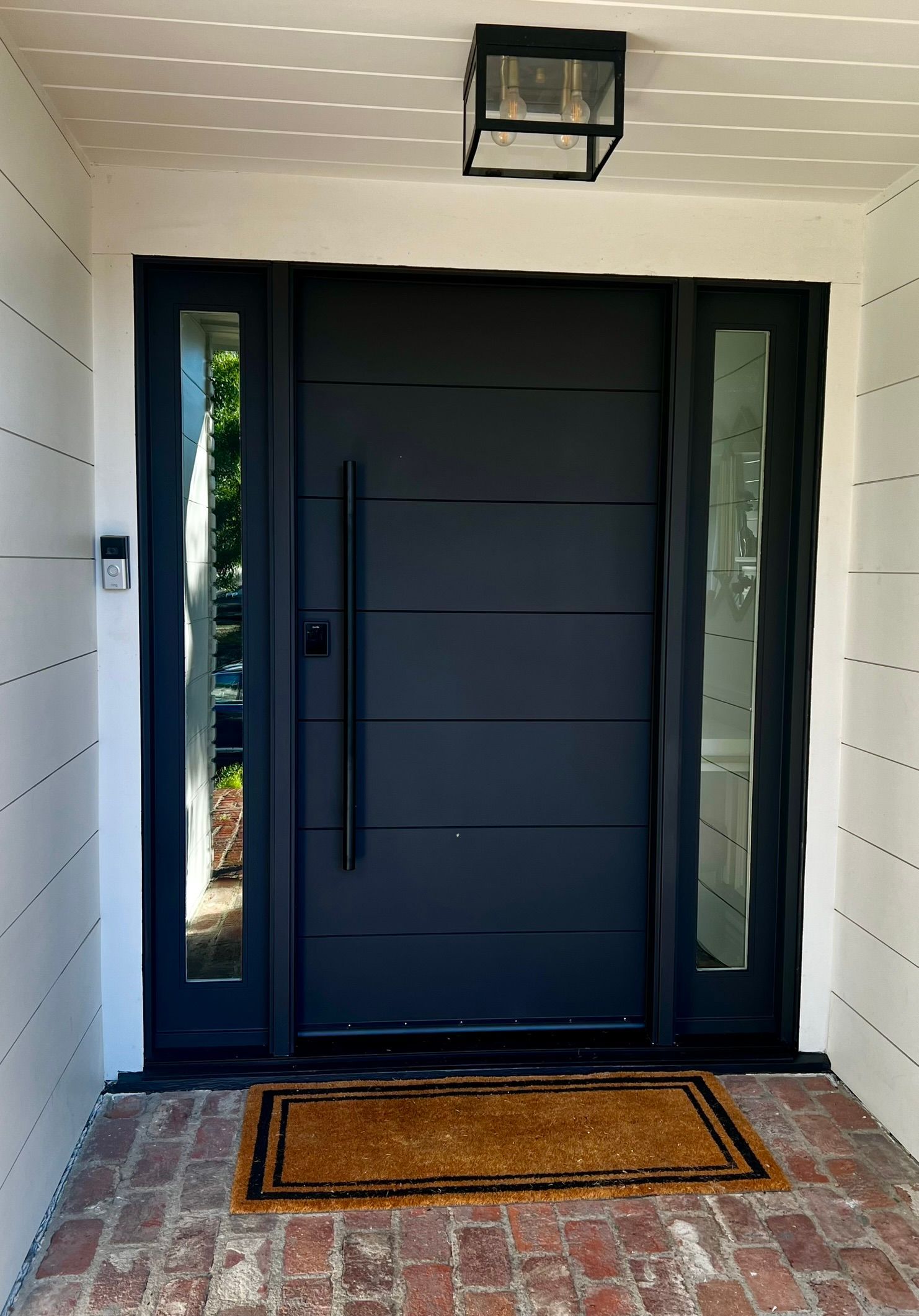 Black front door with sidelights and black hardware; a doormat rests on brick.