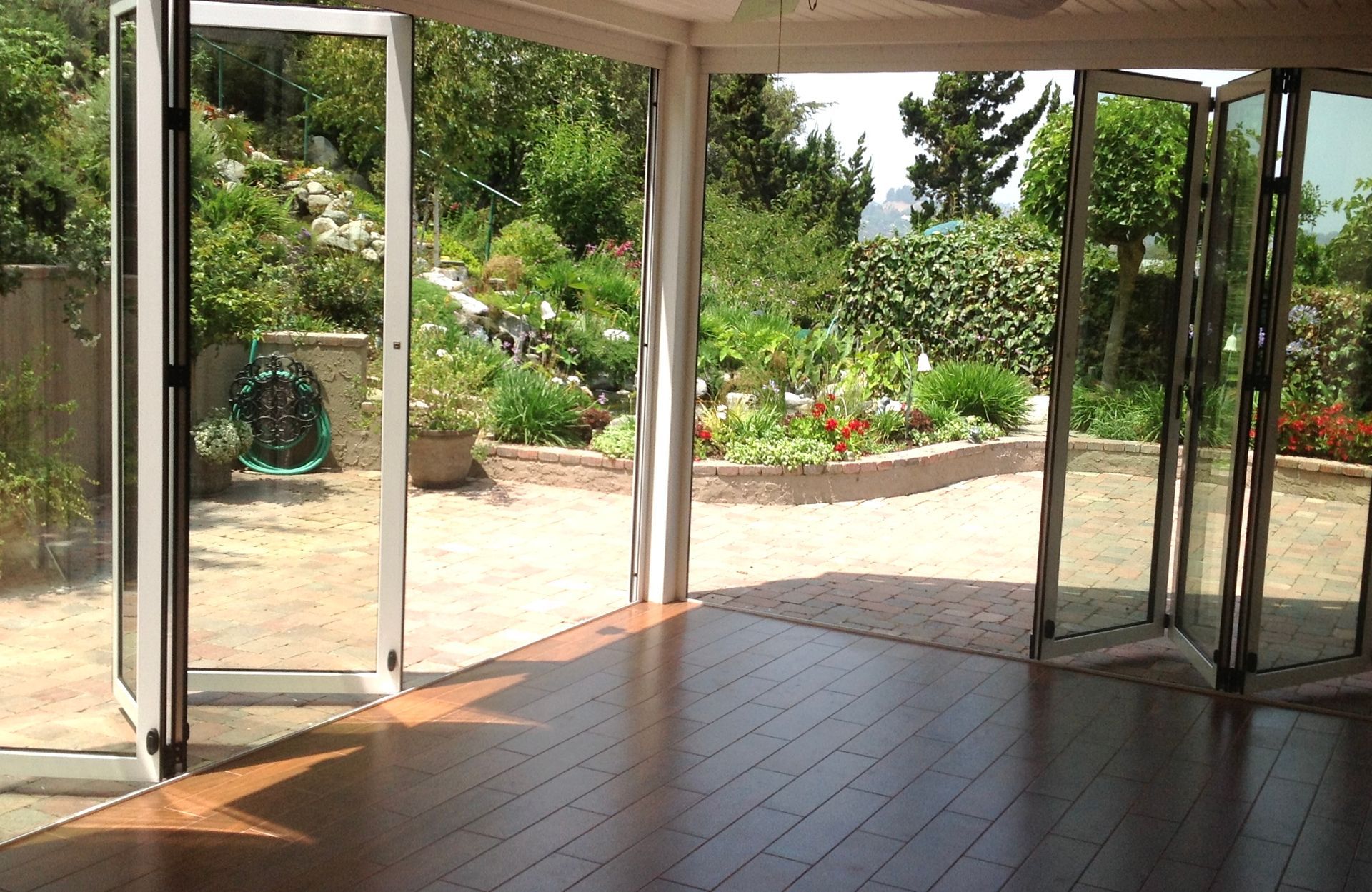 Bifold glass doors open onto a sunny backyard with lush greenery and a brick patio. Wooden floors.