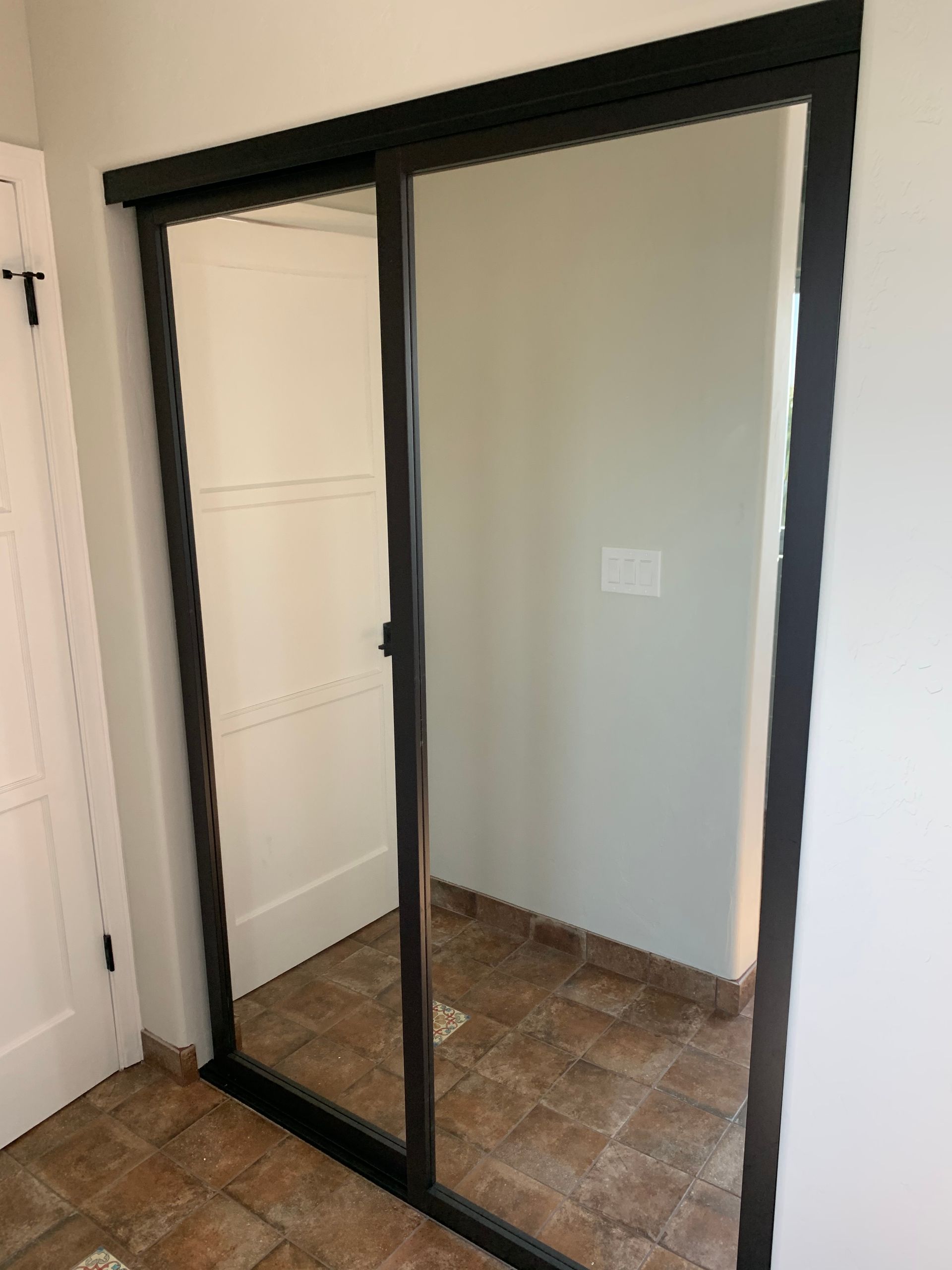 Mirrored sliding closet doors with black frames, reflecting a hallway with tile flooring.