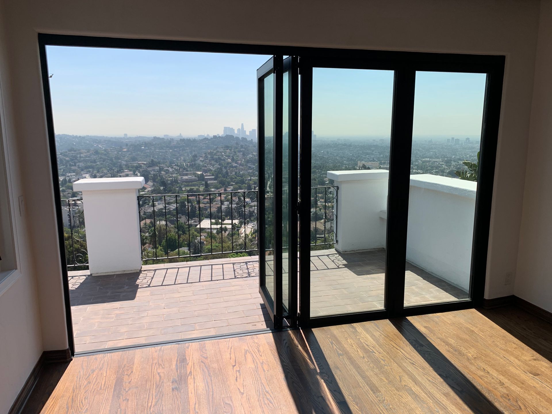 Open doors to a sunny city view. Interior with wood floors, white railings, and a cityscape in the distance.