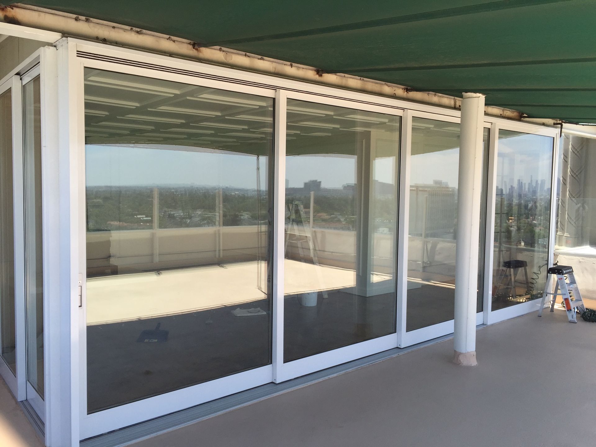 Glass-walled enclosure on a patio with a partial view of a city. White frames, tan floor, green roof.