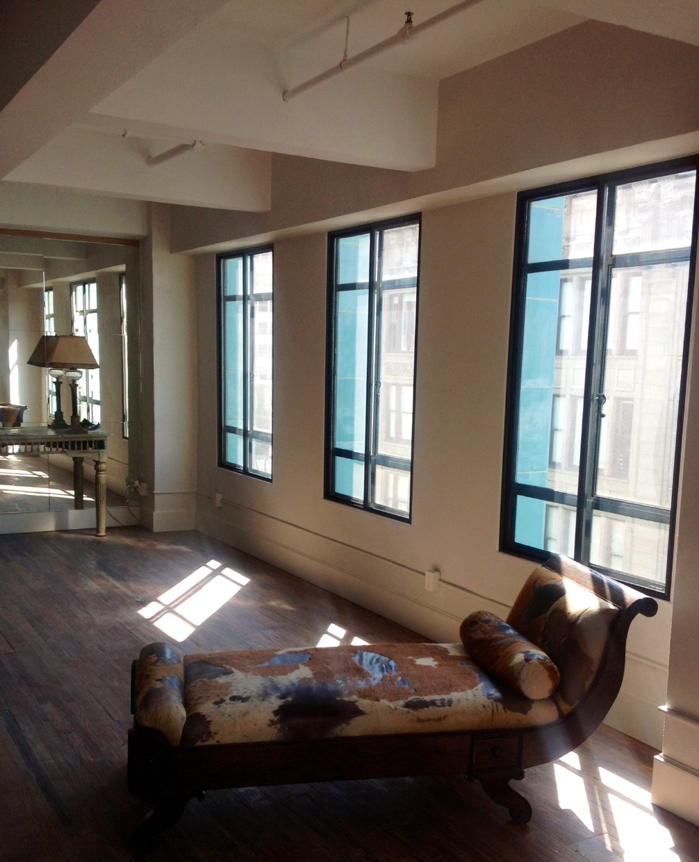 An antique chaise lounge sits in a sunlit, spacious room with large windows and hardwood floors.