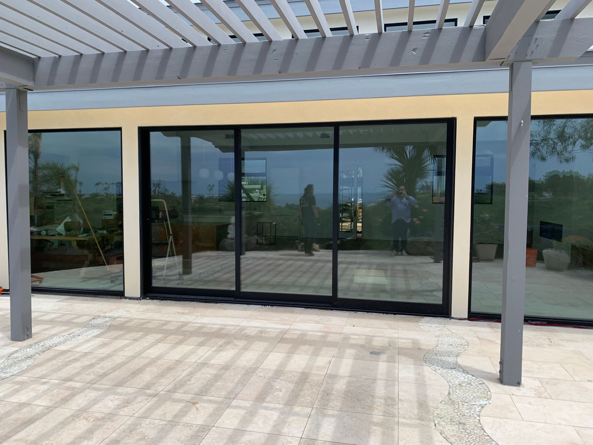 Sliding glass doors with black frames reflecting outdoor scene. Grey pergola, light beige building exterior, stone patio.