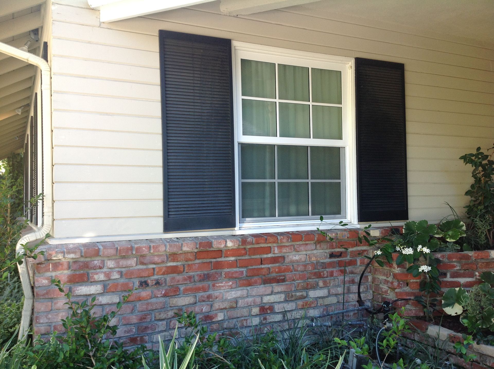 Window with black shutters on a beige house with a brick base, surrounded by greenery.