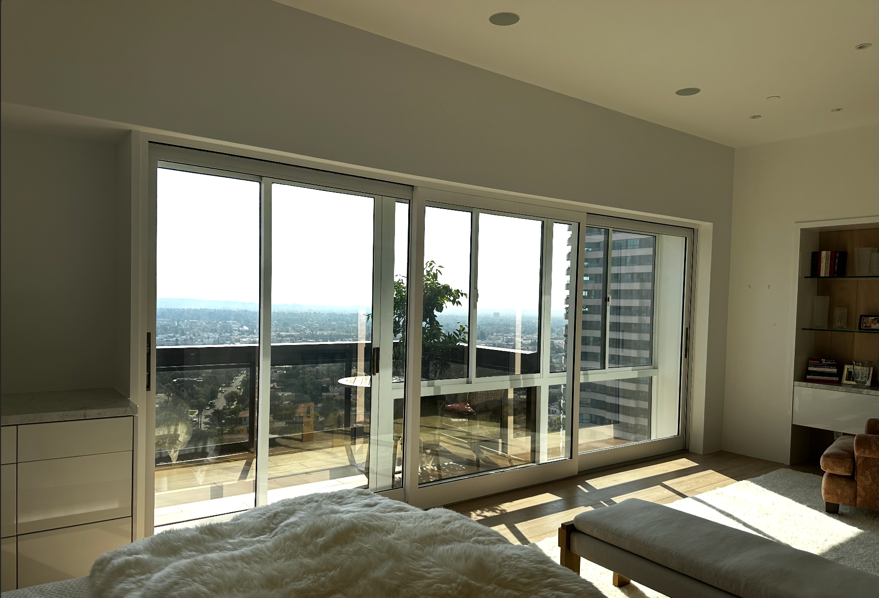 Bright bedroom with large windows overlooking a city skyline. Sunlight streams in.