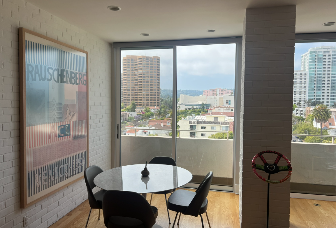 A dining area with a round table, chairs, and cityscape views through a sliding glass door, with a large artwork on the wall.