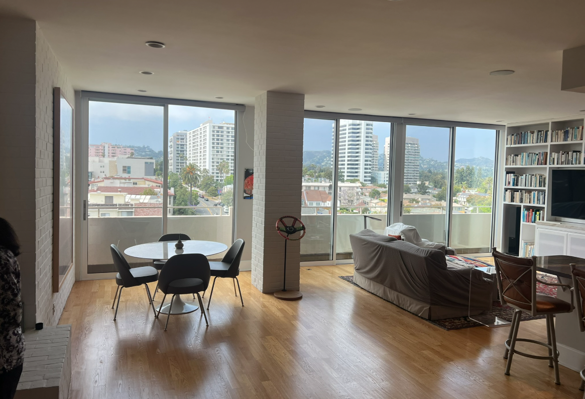 Spacious living area with city view, dining table, sofa, and bookcase. Natural light floods the room.