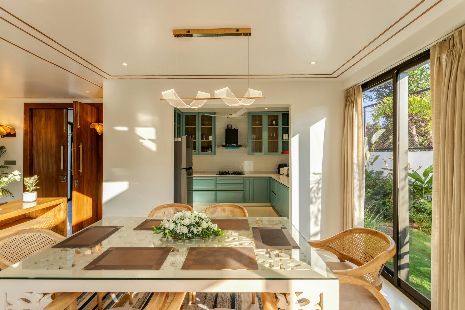 Dining area with a glass table, wicker chairs, and a view of a kitchen and outdoor garden.