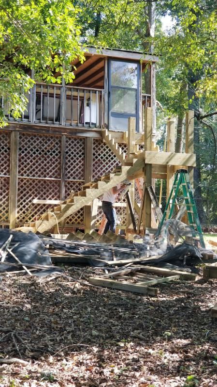 Wooden treehouse under construction with staircase and a person. Green ladder nearby.